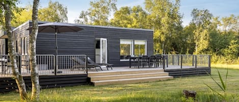 Une cabane moderne aux couleurs sombres, dotée d’une terrasse et d’un espace salon extérieur, entourée d’arbres et d’herbe.