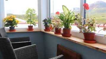 A corner room with a window sill lined with potted plants, wicker chairs, and a view of a road and hills.
