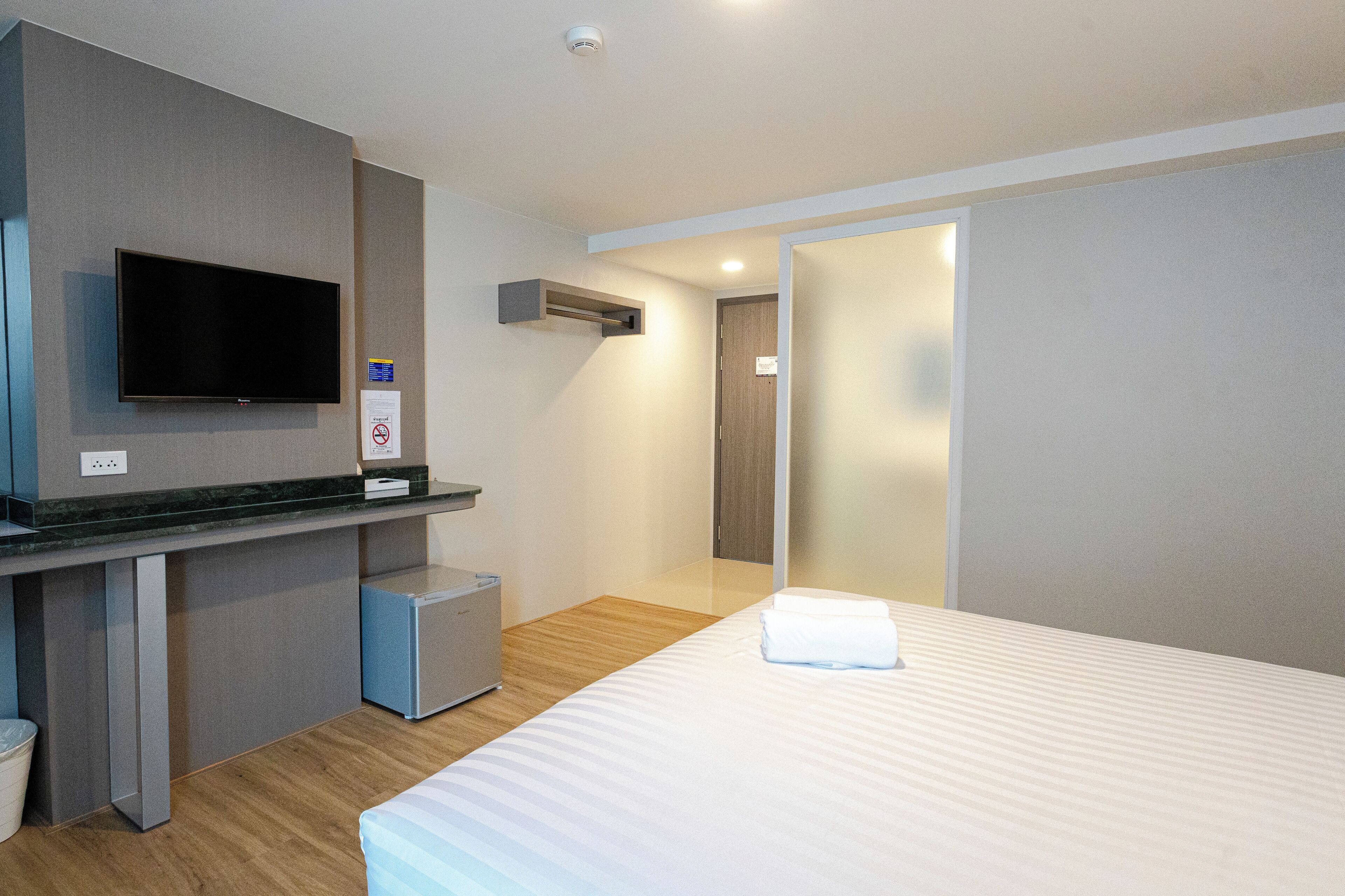 A modern hotel room with a flat-screen TV mounted on the wall, a small desk with a computer, a bed with a neatly folded white towel, and a door leading to another room.