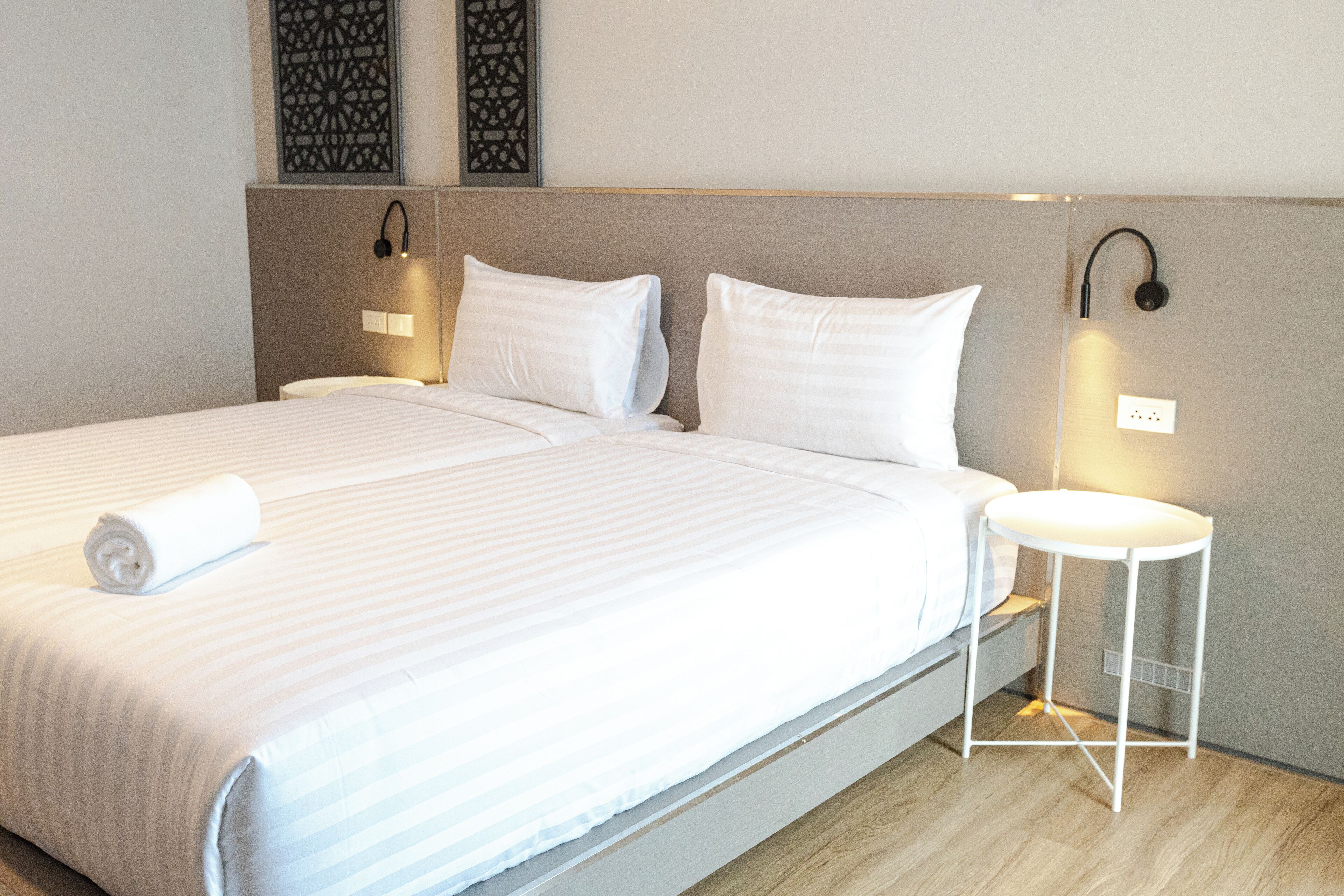Een net opgemaakt bed met witte lakens, een klein nachtkastje met een lamp en wandlampen.