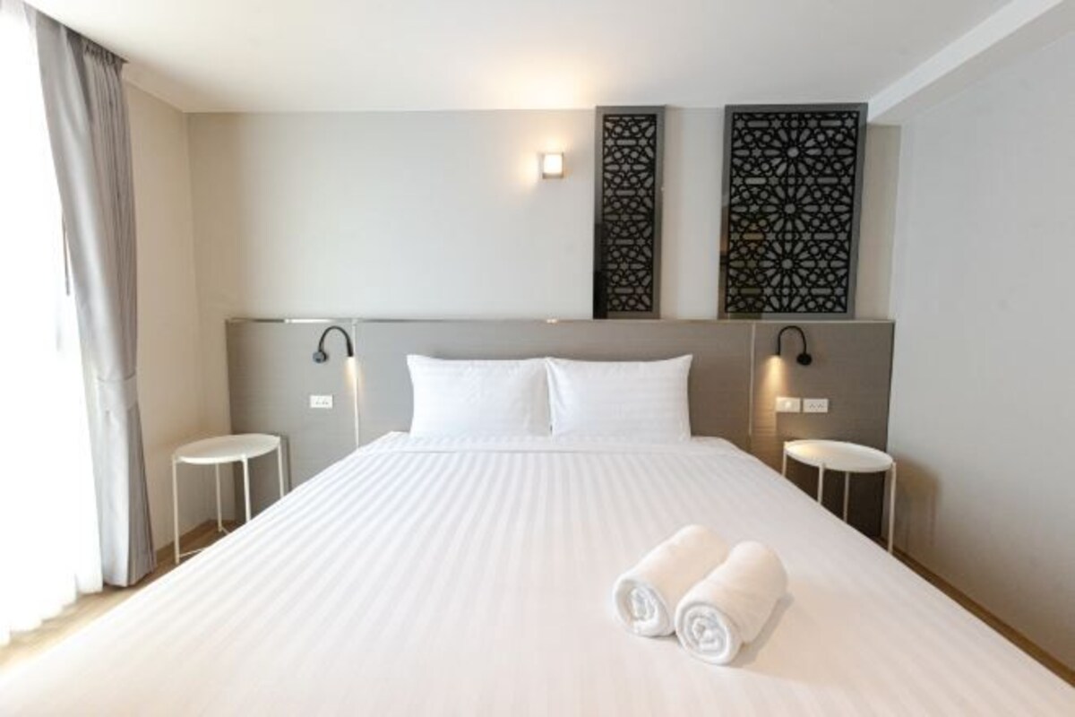Una habitación de hotel con una cama grande, dos mesitas de noche, un cabecero con luces empotradas y paneles decorativos.