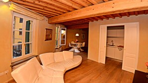 A cozy living room with a curved white sofa, wooden ceiling beams, a dining area with a table and chairs, and a kitchen area with cabinets and a sink.