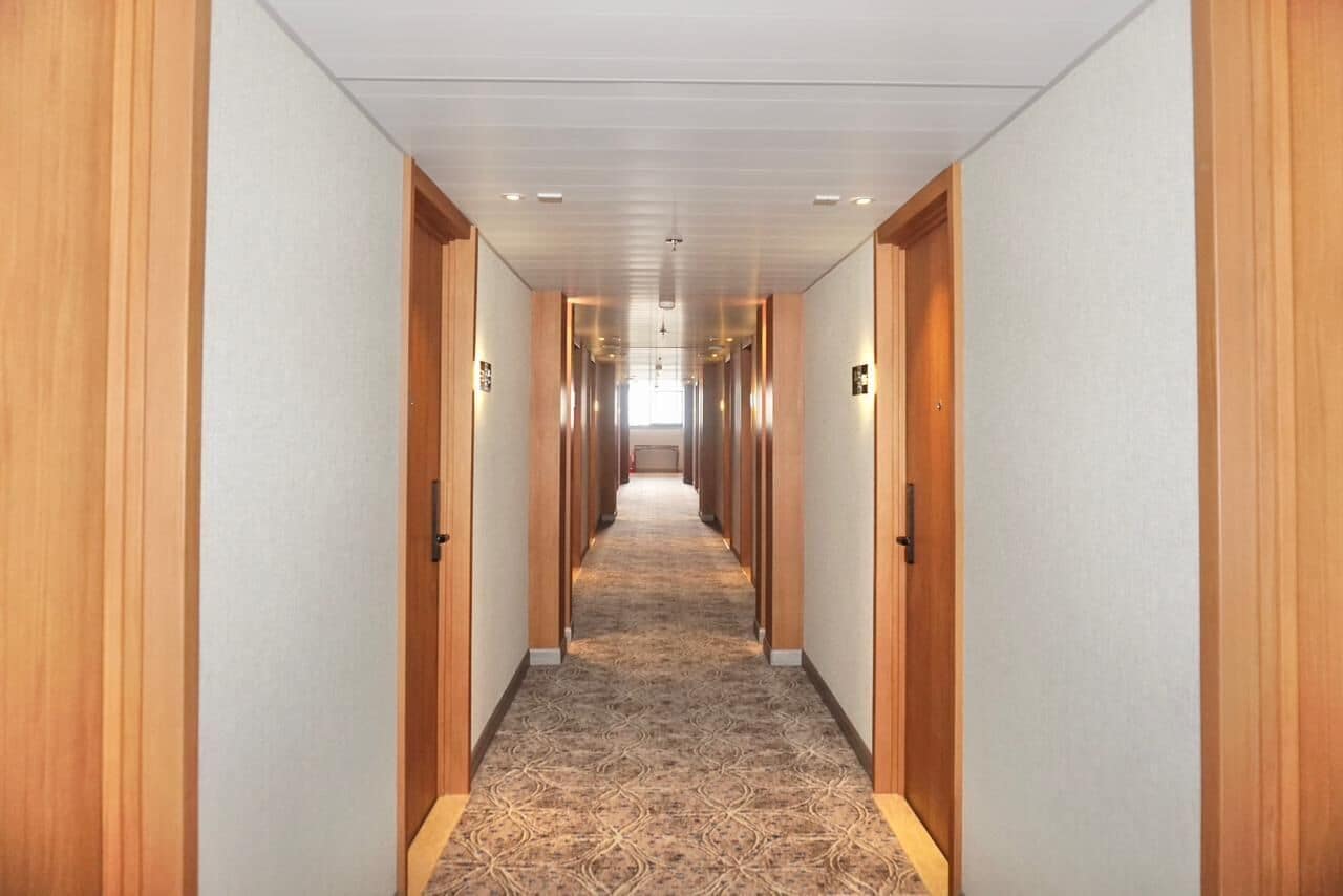 Un pasillo largo y estrecho con puertas de madera a ambos lados, que conduce a una habitación luminosa al final.