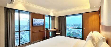 A hotel room with a large bed, a TV mounted on the wall, and a view of the city through large windows.