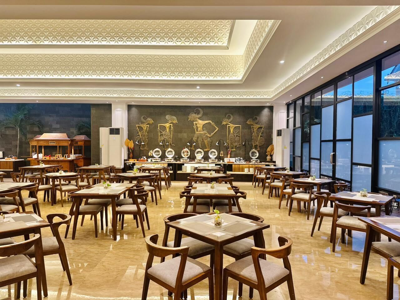 A spacious restaurant with multiple tables and chairs, a decorative wall with sculptures, and large windows providing natural light.