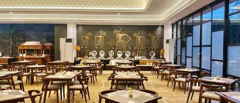 A spacious restaurant with multiple tables and chairs, a decorative wall with sculptures, and large windows providing natural light.