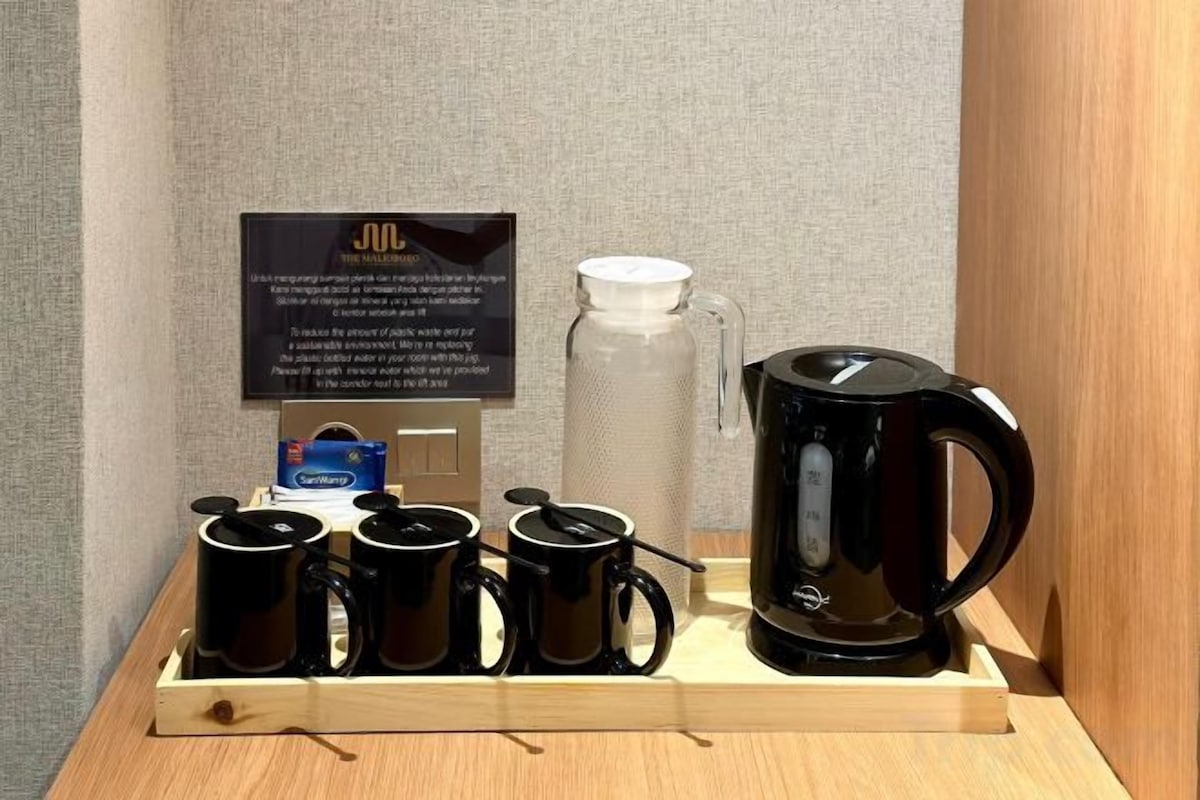 A coffee station with a black coffee maker, four black mugs, a white thermos, and a sign.