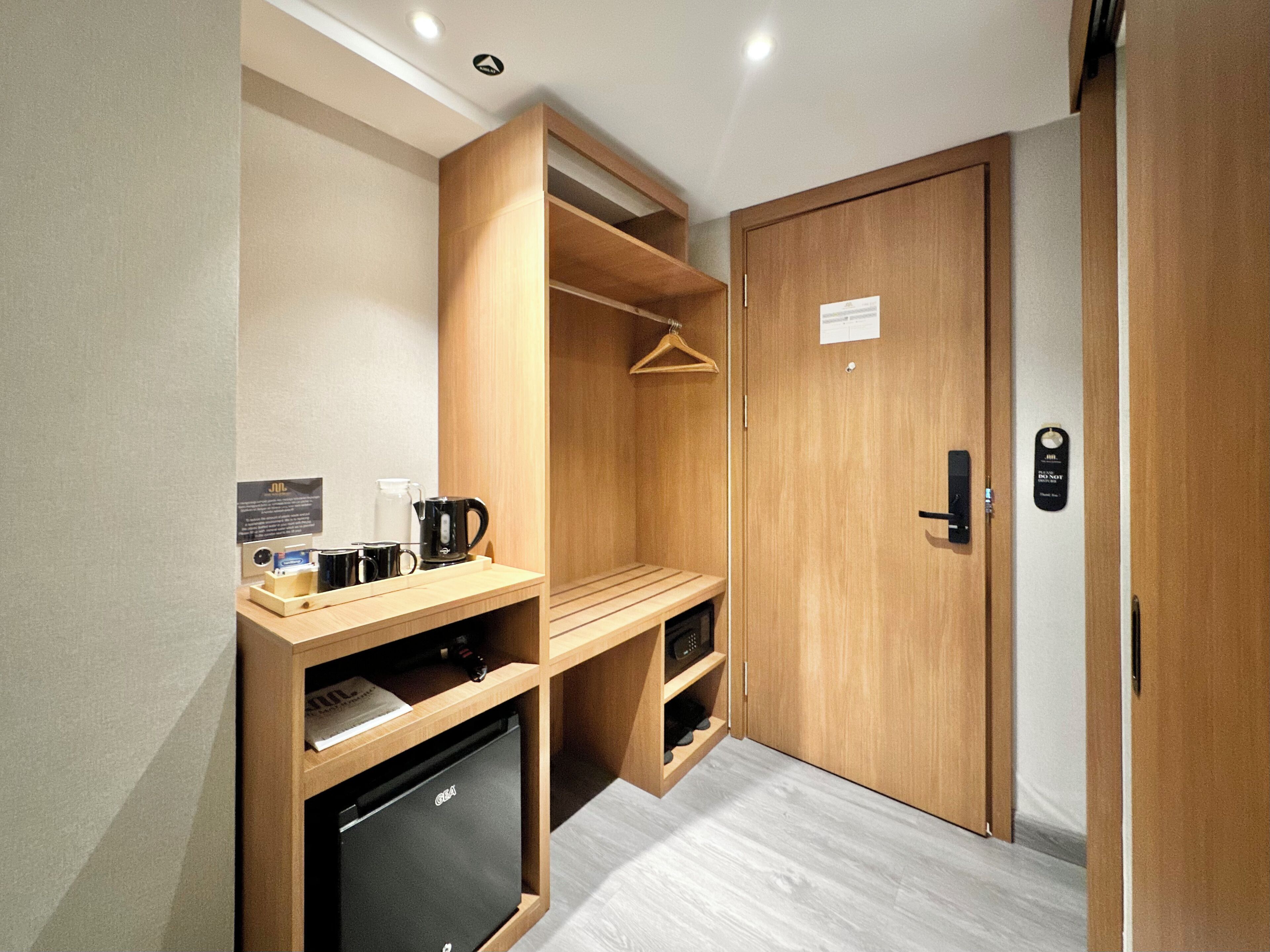A compact room with a wooden wardrobe, a small kitchenette with a refrigerator, a coffee maker, and a hanging clothes rack.
