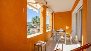 Un balcon avec une table et des chaises, une petite table sur laquelle se trouve un plateau avec des tasses, et une vue sur des palmiers et des immeubles.