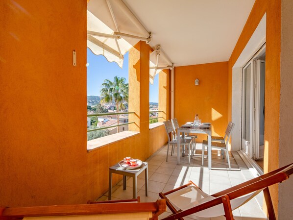 A balcony with a table and chairs, a small table with a tray of cups, and a view of palm trees and buildings.