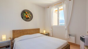 Une chambre à coucher avec une tête de lit en osier, un lit avec une literie blanche, une table de chevet avec une lampe, une assiette décorative accrochée au mur, une fenêtre avec des rideaux blancs et un climatiseur fixé au mur.