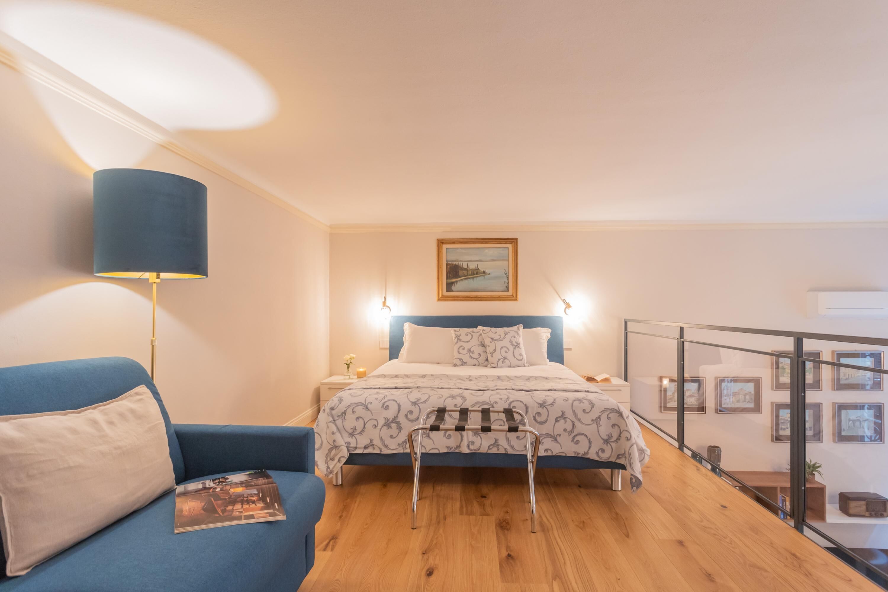 Et soveværelse med seng, blå sofa, et natbord, en lampe og et billede i ramme på væggen.
