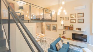 Una sala de estar moderna con un sofá azul, una zona de comedor con una mesa preparada para cuatro personas, y una escalera que sube a un nivel superior.