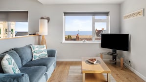 Un salon moderne avec un canapé gris, une table basse en bois, un téléviseur à écran plat et une grande fenêtre donnant sur la mer et les bâtiments.