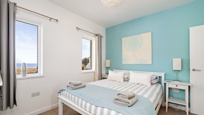 Une chambre à coucher avec un lit, des tables de chevet, une fenêtre avec des rideaux, un tableau au mur et une vue sur la mer.