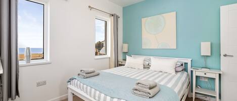 Une chambre à coucher avec un lit, des tables de chevet, une fenêtre avec des rideaux, un tableau au mur et une vue sur la mer.