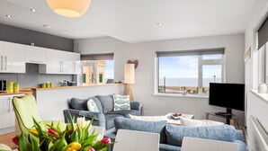 Un salon moderne comprenant un canapé, une table à manger avec des chaises, un téléviseur et une grande fenêtre donnant sur la mer.