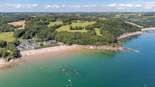 Vue aérienne d’une zone côtière comprenant une plage, un parking et un vaste domaine entouré de verdure.