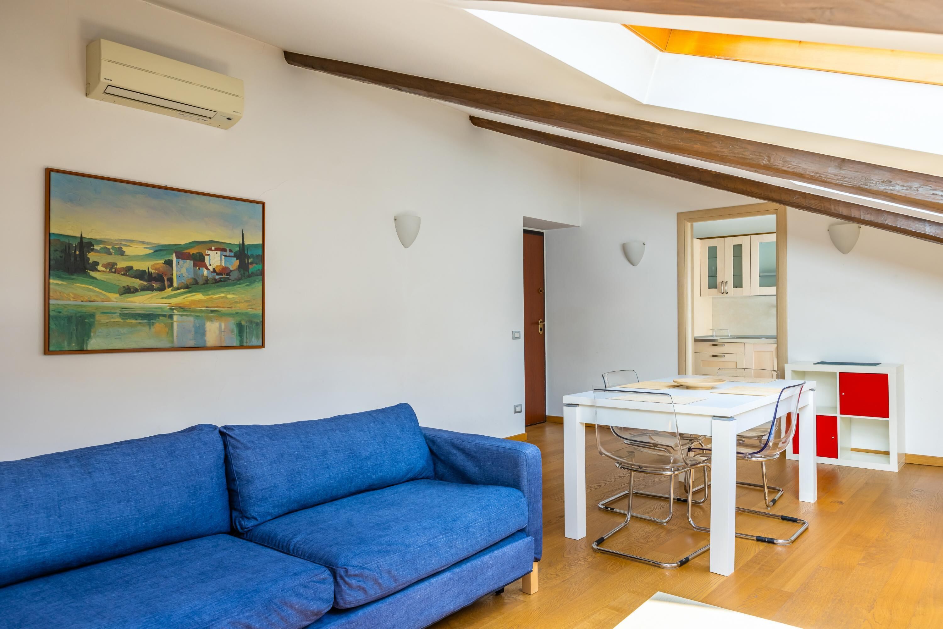 A modern living room with a blue sofa, a dining table with chairs, a kitchen area, and a skylight.