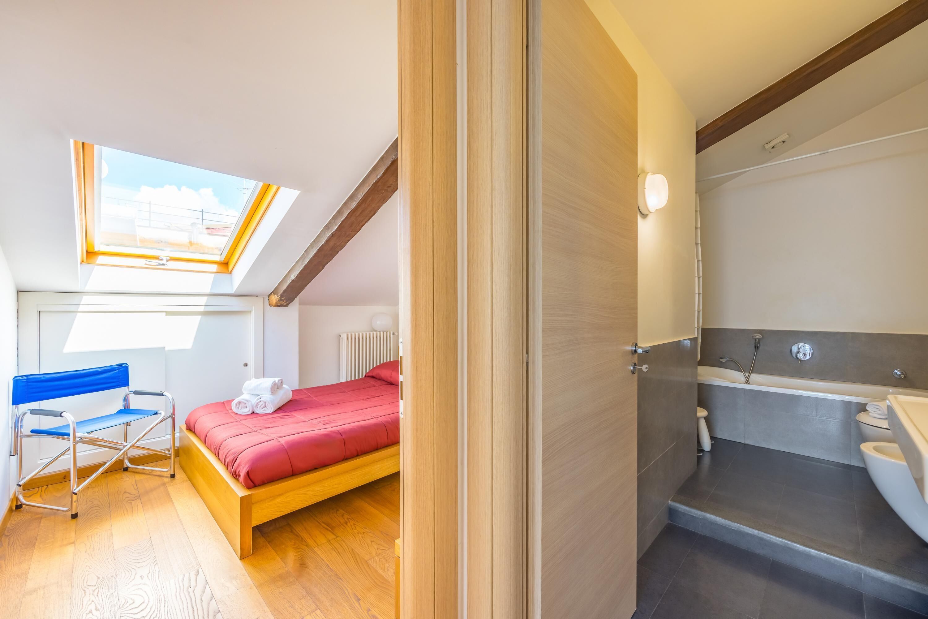 A bedroom with a skylight, a bed, a chair, and a bathroom with a bathtub and toilet.