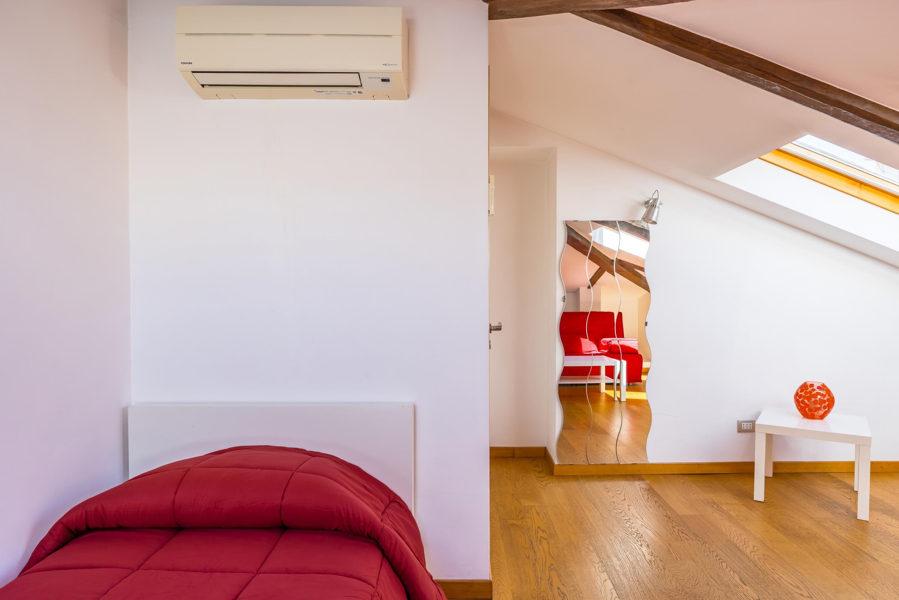 A bedroom with a red bed, a white wall, an air conditioning unit, a wooden floor, a white table with a red vase, and a skylight.