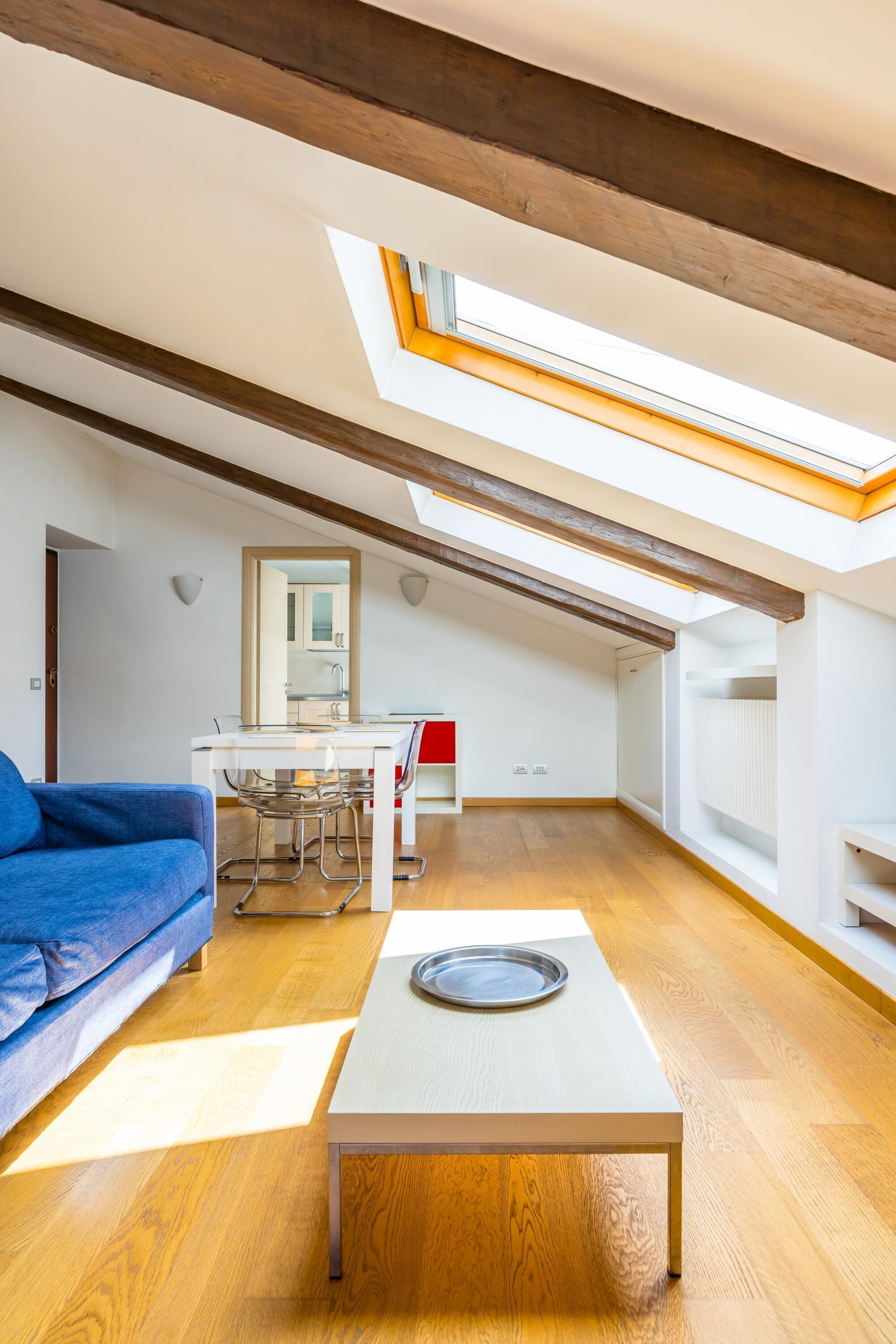 A modern loft apartment with a skylight, wooden beams, a dining area, and a blue sofa.