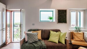 Uma sala de estar com um sofá marrom, almofadas verdes e amarelas, um cobertor estampado e uma janela com vista para o exterior.