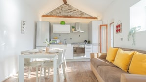 Cozinha compacta com armários brancos, área de refeições com mesa e cadeiras brancas, e um sofá marrom com almofadas amarelas.
