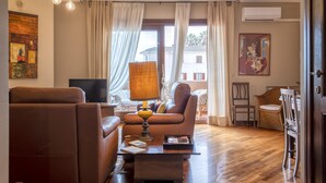 Een woonkamer met een bruine bank, een salontafel, een lamp, een televisie en een eethoek met rieten stoelen.