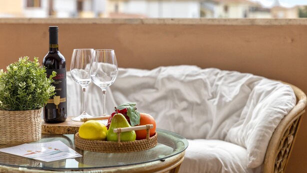 Een glazen tafel met een fles wijn, twee glazen, een mand met fruit en een kleine plant op een balkon met uitzicht op gebouwen.