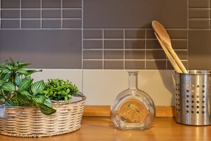 Una encimera de cocina con una cesta de mimbre que contiene plantas verdes, una botella de vidrio transparente con etiqueta y un portalápices metálico con utensilios de madera.