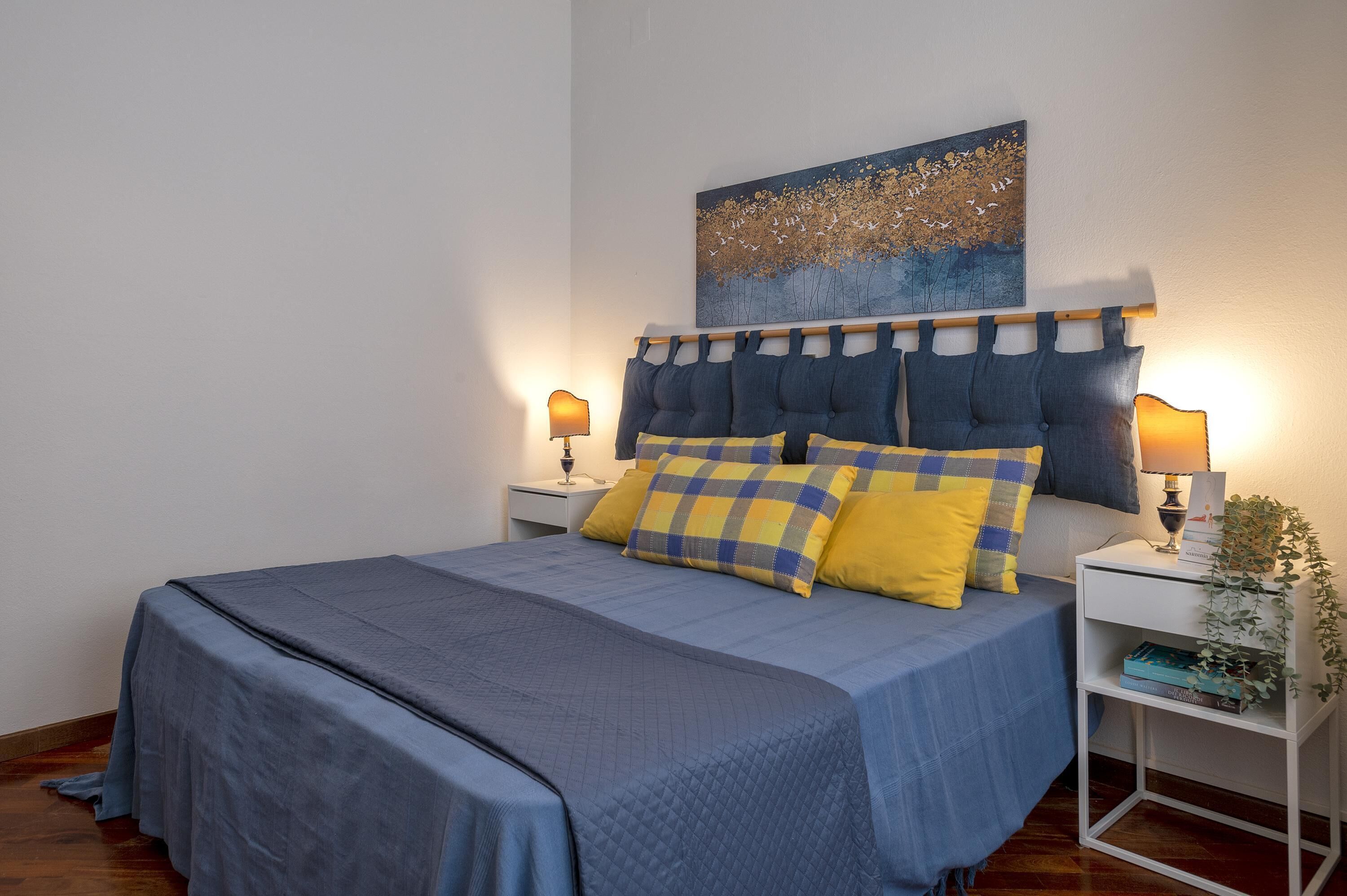 A neatly made bed with a blue bedspread and two pillows, one yellow and one blue. There is a wall-mounted artwork above the bed and a small white nightstand with a lamp beside it.