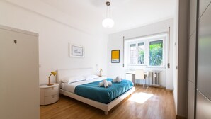 Quarto moderno com cama, mesa de cabeceira, abajur e janela com vista para o verde.