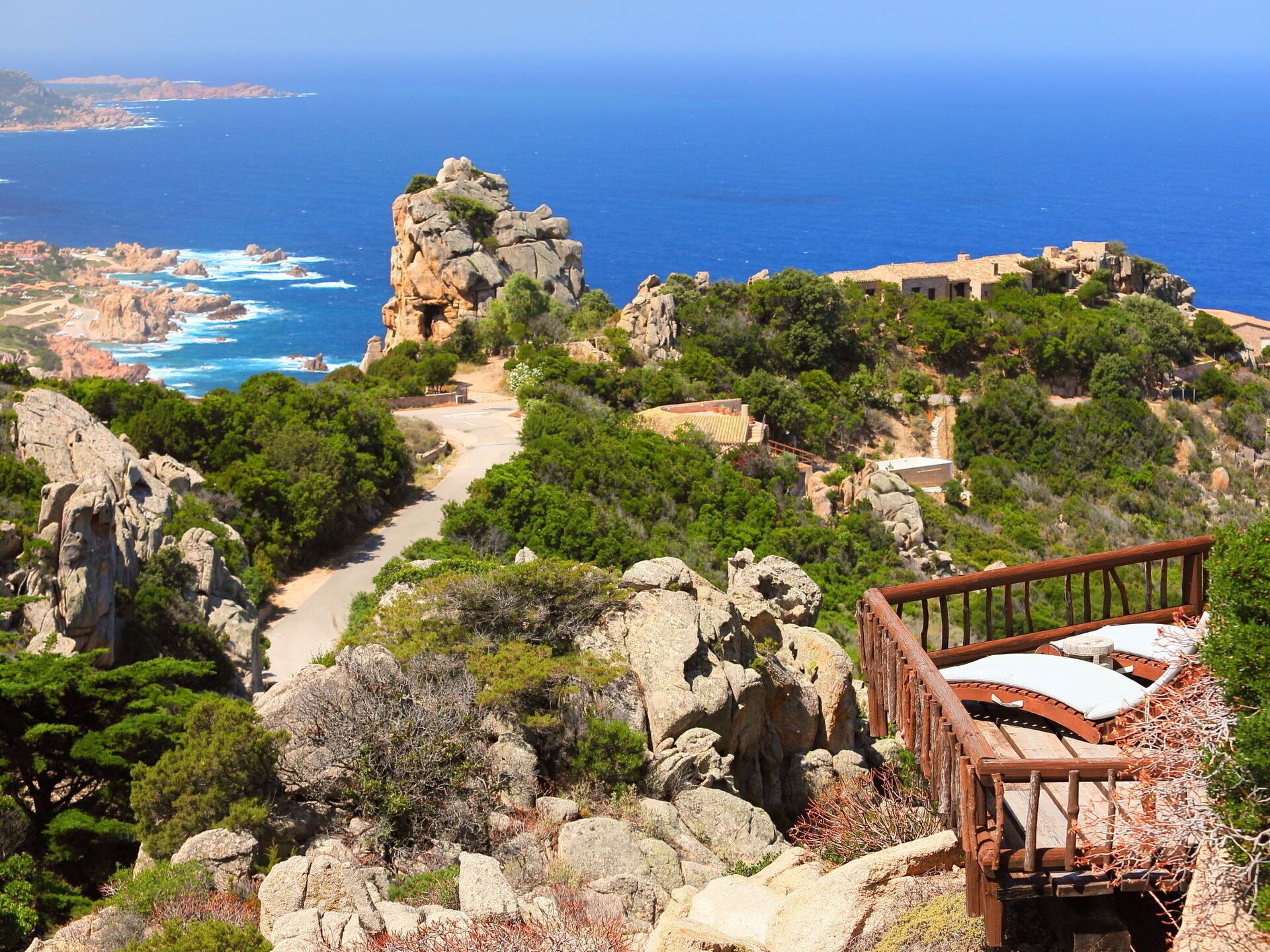 A coastal landscape with a rocky outcrop, a wooden viewing platform, and a path leading to a building with a tiled roof.