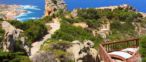 A coastal landscape with a rocky outcrop, a wooden viewing platform, and a path leading to a building with a tiled roof.