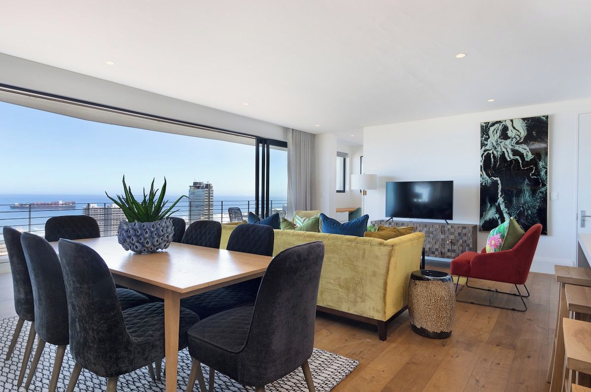 Un salon moderne comprenant un coin repas, un canapé, un téléviseur et une grande fenêtre donnant sur la mer.
