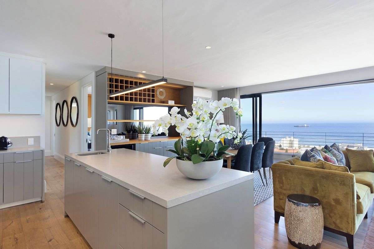 A modern kitchen with a central island, a dining area with a yellow sofa, and a view of the sea.