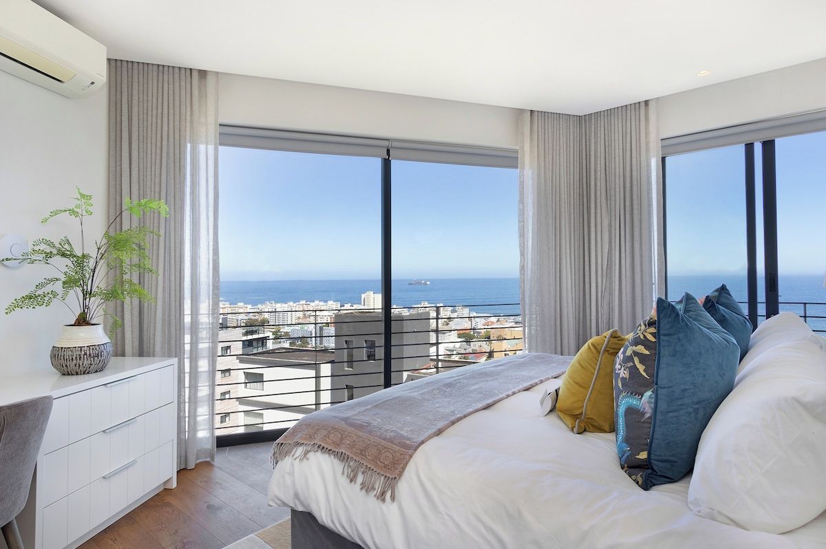 A bedroom with a large bed, a bedside table, a potted plant, and a view of the city and sea through floor-to-ceiling windows.