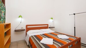 Een eenpersoonsbed met een gestreept laken, twee nachtkastjes met lampen, een houten plank en een radiator.