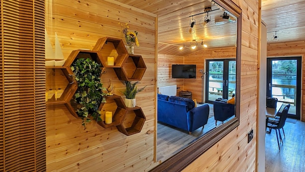 A wooden interior with a large mirror, hexagonal shelves with candles, and a blue sofa in the background. - Ferry Lodge - Norfolk Broads (Brundall)