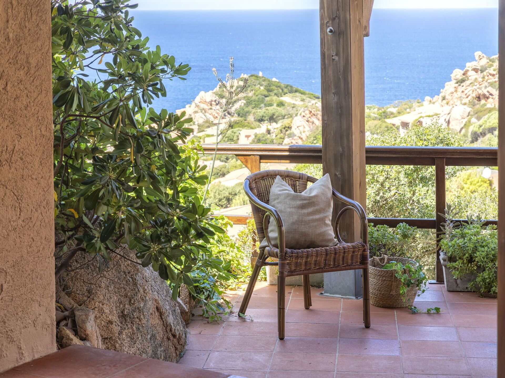 Une chaise en osier avec un coussin, placée sur un balcon donnant sur un paysage rocheux et la mer.