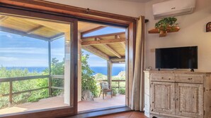 Habitación con puerta de vidrio corredera que da a un balcón con vista al mar. Hay un mueble de madera con un televisor encima y un equipo de aire acondicionado en la pared.