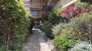 A narrow stone pathway flanked by lush greenery and vibrant flowers leading to a building with multiple balconies.