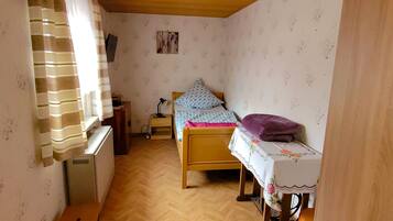 A small, compact room with a single bed, a nightstand, a desk, and a radiator. The room has a wooden ceiling and walls with floral wallpaper.