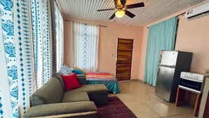 Un espacio habitable compacto con sofá, cama con ropa de cama estampada, ventilador de techo, unidad de aire acondicionado y una pequeña kitchenette.