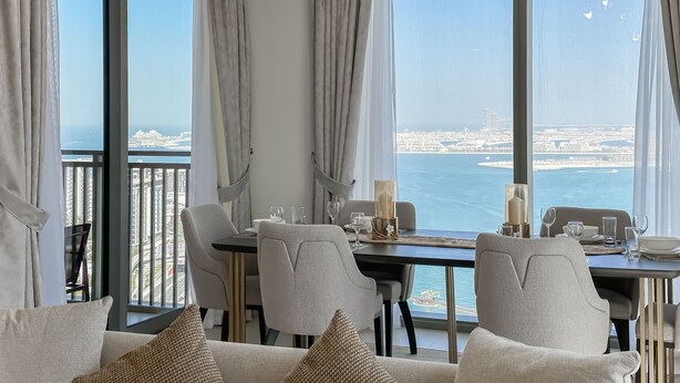 A dining area with a table set for four, large windows with curtains, and a view of the sea and cityscape.