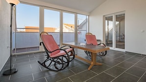 Ein Balkon mit zwei Schaukelstühlen, einem Tisch, großen Fenstern und einer Stehlampe.