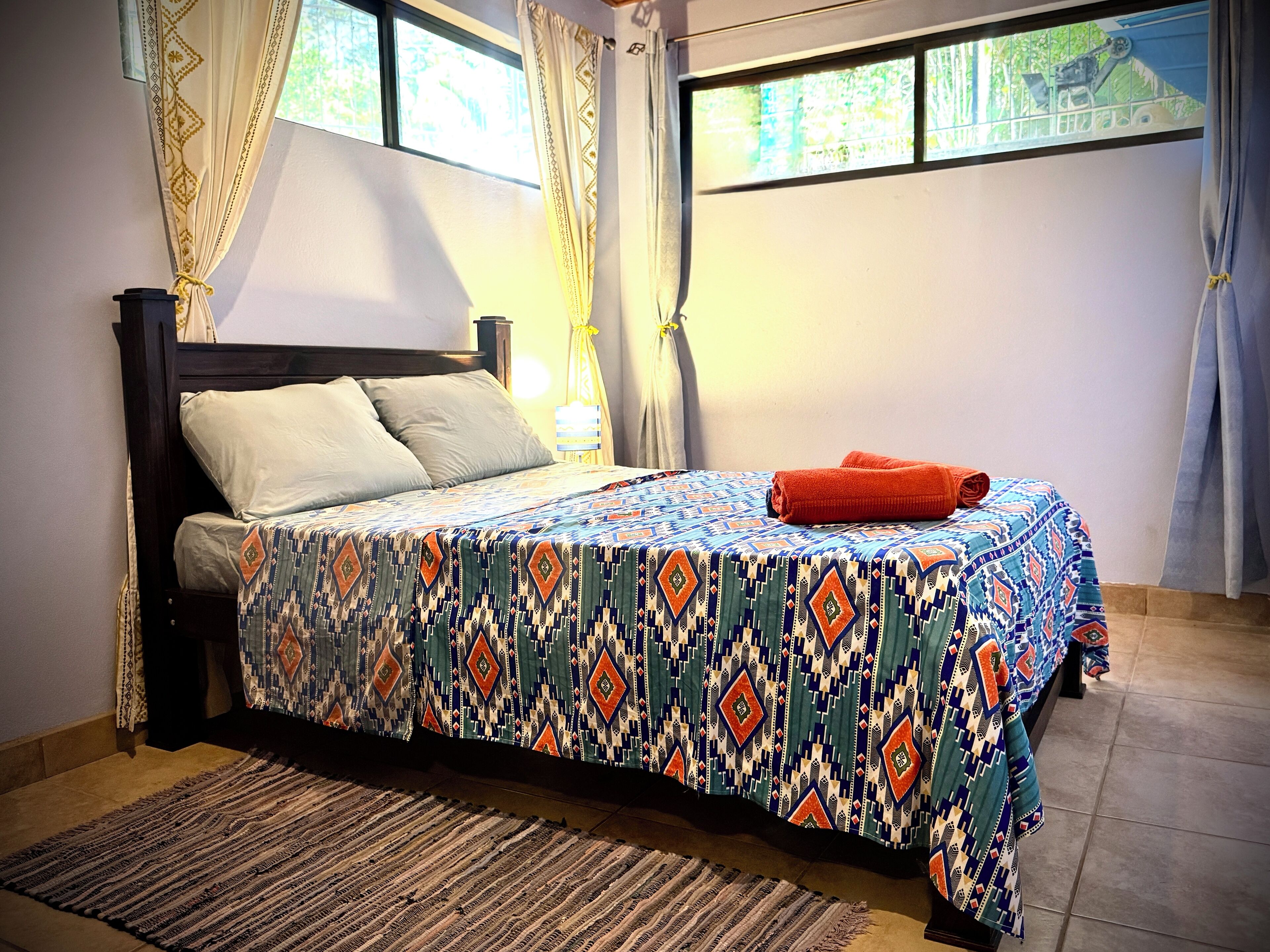 Un lit bien fait, recouvert d’une couvre-lit à motifs, une lampe de chevet et une fenêtre avec des rideaux.