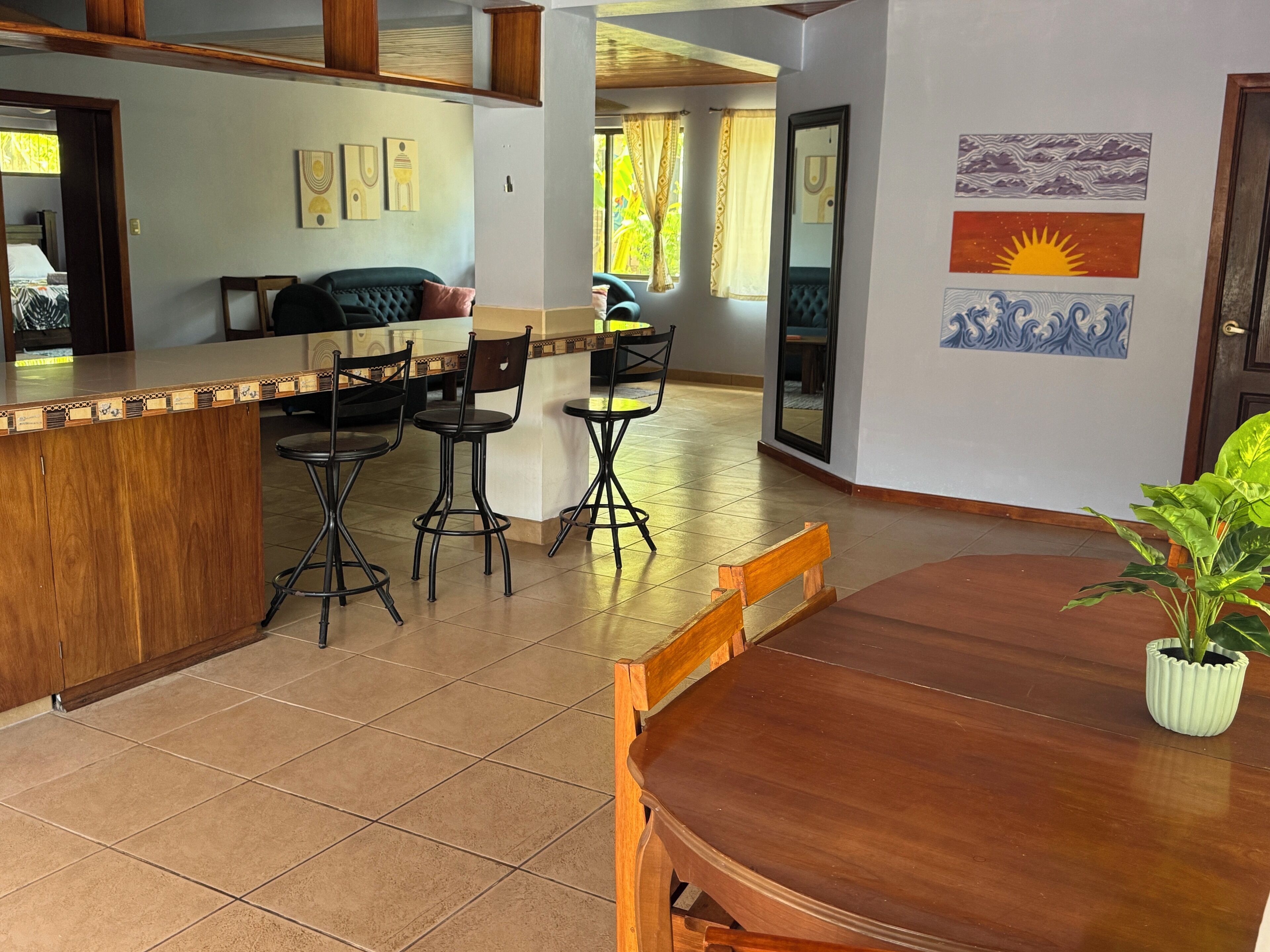 Cozinha espaçosa com bancada e área de bar em madeira, equipada com banquetas. Há uma mesa de jantar com cadeiras e um vaso de planta.