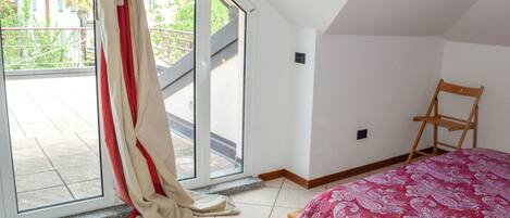 A bedroom with a bed, a chair, a sliding glass door, and a view of a balcony with a railing.